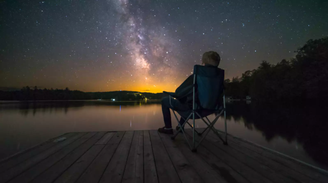 Mensch in Campingstuhl auf einem Steg am See, mit dem Rücken zum Betrachter schaut in den Sternenhimmel.