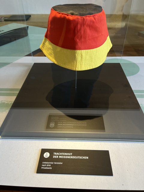 Ein Schlapphut in den Farben schwarz-rot-gelb in einer Ausstellungsvitrine. Beschriftung auf der Plakette davor: „TRACHTENHUT DER MEISSNERDEUTSCHEN Unbekannter Hersteller nach 2018 Privatbesitz“