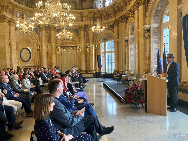 Man sieht einen pompösen Festsaal mit einem Kronleuchter an der Decke, er befindet sich im Neuen Schloss in Stuttgart. Rechts im Bild sieht man Minister Manne Lucha. Er steht an einem Podium und spricht zu einem Publikum. Links im Bild sieht man ein Publikum, das auf Stühlen sitzt. Es handelt sich um den Iftar-Empfang der Landesregierung.