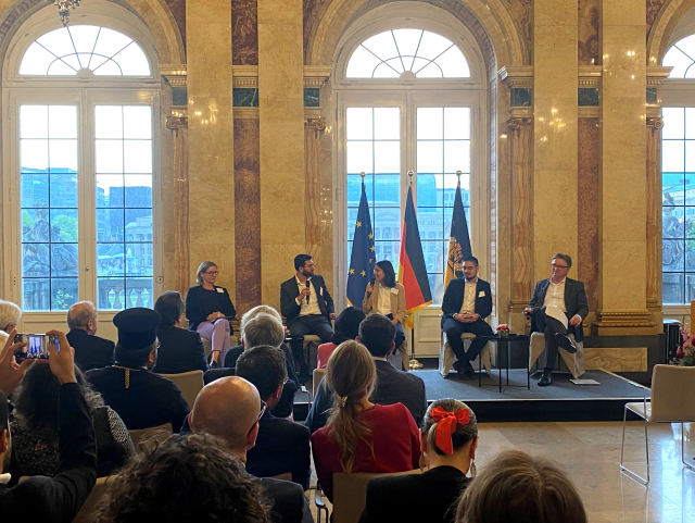 Das Foto zeigt fünf Personen, die in einem Raum vorne auf einem Podium sitzen. Sie befinden sich im Neuen Schloss in Stuttgart. Rechts auf dem Podium sitzt Minister Manne Lucha. Ein Mann auf dem Podium spricht gerade. Vor dem Podium sitzen viele Menschen in einem Publikum. Hinter dem Podium befinden sich eine EU-Flagge, eine Deutschland- und eine Baden-Württemberg-Flagge. Es handelt sich um den Iftar-Empfang der Landesregierung.