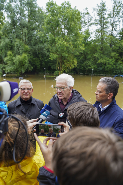 Von links nach rechts: Thomas Strobl (Innenminister von Baden-Württemberg), Winfried Kretschmann (Ministerpräsident von Baden-Württemberg) und Georg Schellinger (Bürgermeister von Meckenbeuren) geben vor dem überfluteten Schulhof in Kehlen ein Interview. 