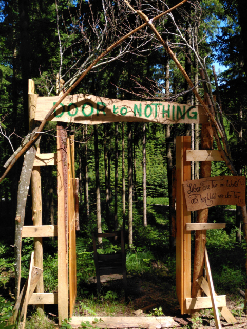 Ein aus Bettern und Ästen gezimmerter Türrahmen steht im Wald. Durch die Öffnung blickt man in eine Senke und damit tief in den Wald. Über der Tür steht in grüner Farbe handgemahlt „Door to Nothing“.