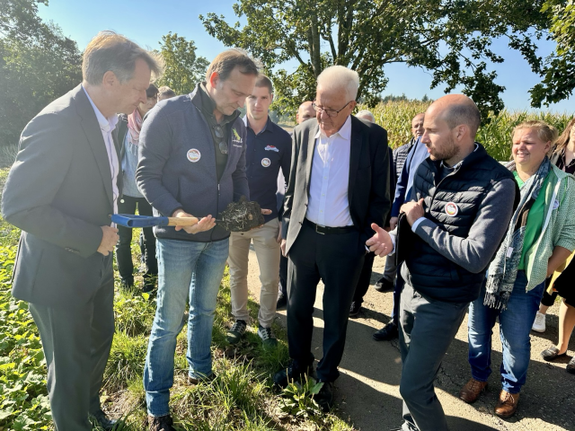 Ministerpräsident Kretschmann steht mit Vertreterinnen und Vertretern der Bürgerinitiative Waldhof am Rand eines Kartoffelfeldes und lässt sich den besonderen Erdboden zeigen. Die Erde wird ihm auf einer Schaufel hingehalten.