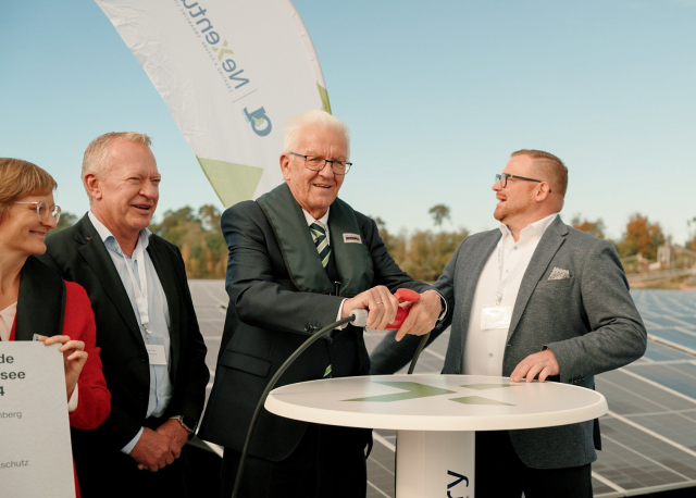 Ministerpräsident Kretschmann steht an einem Tisch und nimmt mit einem Stecker die Solaranlage in Betrieb. Hinter ihm sieht man die Solarzellen.