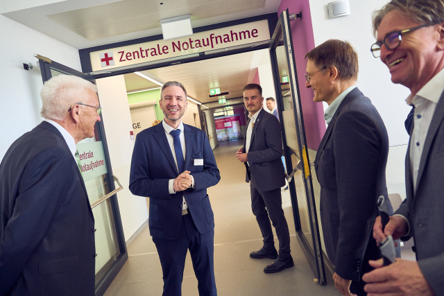 Ministerpräsident Kretschmann steht mit Chefarzt Dr. Heiner Stäudle, Bundesgesundheitsminister Lauterbach und BW-Gesundheitsminister Manne Lucha vor dem Eingang der Zentralen Notaufnahme. Alle vier lachen. Im Hintergrund ist der Krankenhaus-Flur zu sehen. 