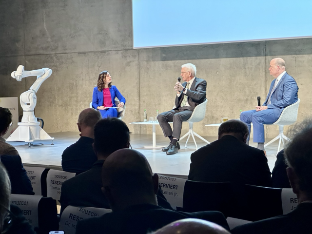 Seitlicher Blick von hinten aus dem Publikum auf die Bühne, wo die Moderatorin (links) mit Ministerpräsident Kretschmann (Mitte) und einem weiteren Panel-Teilnehmer sitzt. Der Ministerpräsident spricht in sein Mikrofon. 