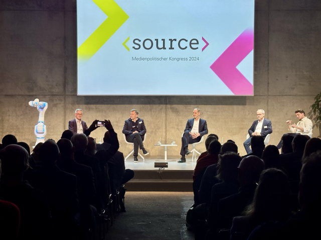 Blick von hinten aus dem Publikum auf die Bühne, wo die Moderator (rechts) mit vier weiteren männlichen Gästen sitzt. Hinter ihnen ist ein Bildschirm mit dem Schriftzug „Source - Medienpolitischer Kongress 2024“ zu sehen.