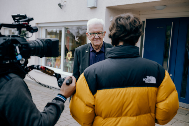 Ministerpräsident Kretschmann schaut zu dem Mann in gelb-schwarzer Ballonjacke, der in interviewt. Rechts neben dem Mann ist ein zweiter Mann mit Kamera auf der Schulter. Im Hintergrund die Grundschule. 