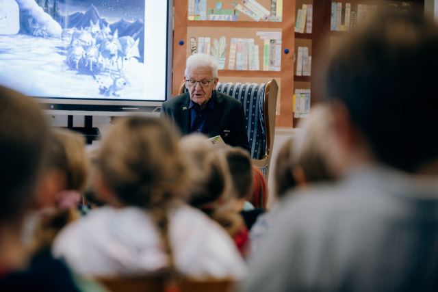 Ministerpräsident Kretschmann sitzt in einem blaugestreiften Ohrensessel und schaut lesend nach unten. Im Vordergrund Umrisse der Kinder, im Hintergrund eine Tapete mit Büchern.