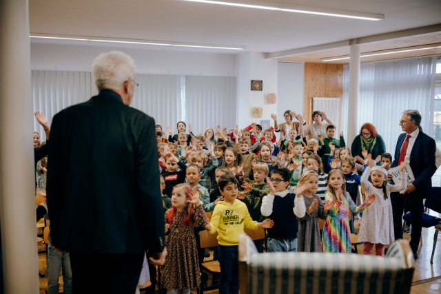 Ministerpräsident Kretschmann ist von hinten zu sehen. Vor ihm stehen die Schülerinnen und Schüler. Sie singen, klatschen und heben die Arme.