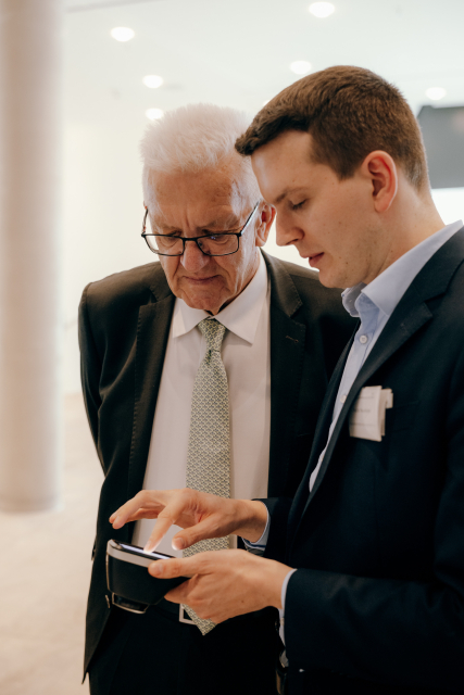 Ministerpräsident Kretschmann steht neben einem Mann, der ihm auf einem kleinen digitalen Gerät etwas zeigt. Beide schauen nach unten.