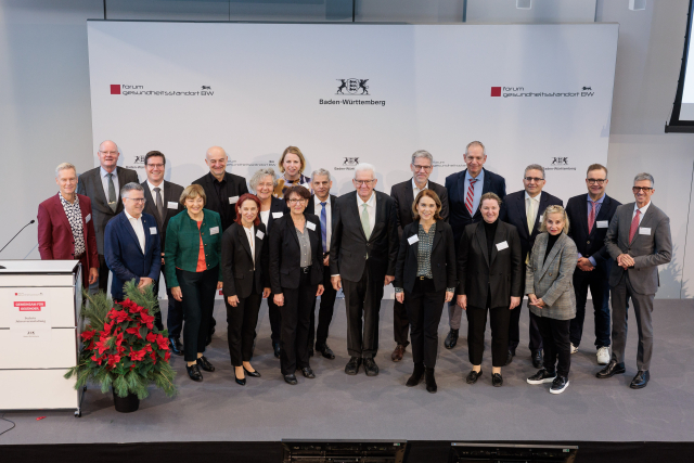 Zwanzig Menschen stehen nebeneinander auf einer Bühne. Alle sind schick gekleidet. In der Mitte steht Ministerpräsident Kretschmann neben Ministerin Petra Olschowski. Rechts neben ihm stehen Staatssekretärin @Dr. Ute Leidig und Staatssekretär Dr. @Patrick Rapp. Auf dem weißen Hintergrund ist das Wappen von Baden-Württemberg aufgedruckt.