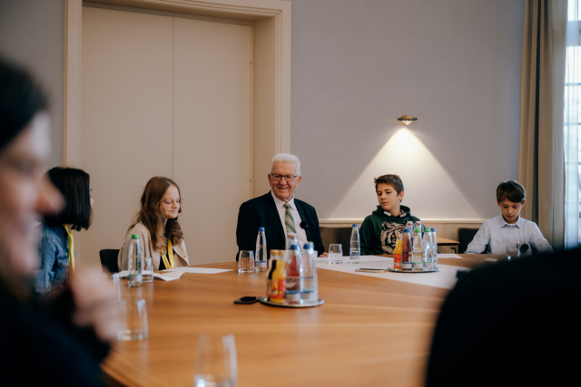 Ministerpräsident Winfried Kretschmann sitzt mit vier Kinder-Reportern am runden Tisch im Kabinettsaal der Villa Reitzenstein. Links von ihm sitzen zwei Mädchen, auf seiner rechten Seite zwei Jungen. © Staatsministerium BW | Ilkay Karakurt