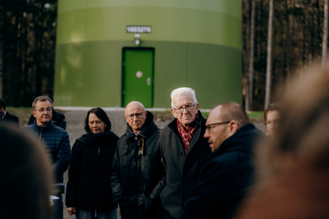 Ministerpräsident Winfried Kretschmann steht in einer Gruppe von Menschen. Im Hintergrund sind Teile des Sockels und des grünen Turms eines Windrads zu sehen. © Staatsministerium BW | Ilkay Karakurt