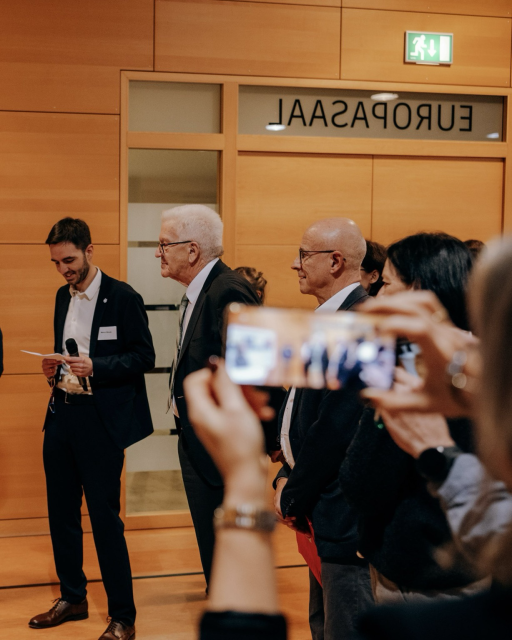 Ministerpräsident Winfried Kretschmann hört gemeinsam mit anderen Personen einem Vortrag zu und wird dabei mit Handy fotografiert. Foto: Staatsministerium BW | Ilkay Karakurt