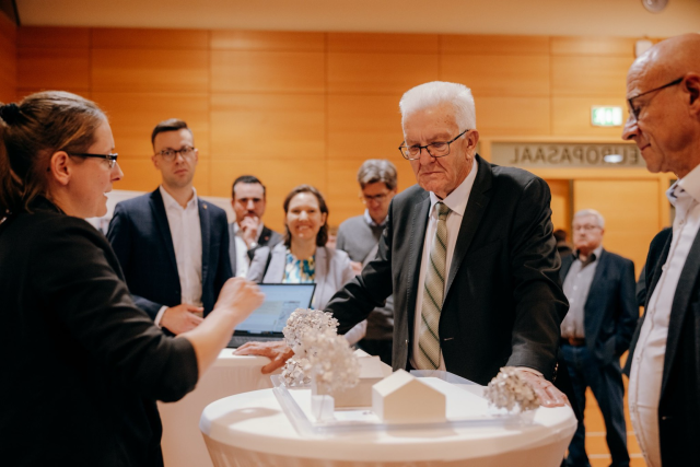 Ministerpräsident Kretschmann betrachtet ein Modell, das ihm von einer Frau erklärt wird. Im Hintergrund sind weiter Personen zu sehen. Foto: Staatsministerium BW | Ilkay Karakurt