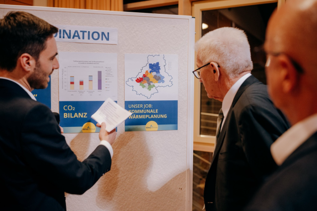 Ministerpräsident Winfried Kretschmann betrachtet gemeinsam mit einem Mann eine Infotafel mit Plakaten zur CO2-Bilanz und zur kommunalen Wärmeplanung. Foto: Staatsministerium BW | Ilkay Karakurt