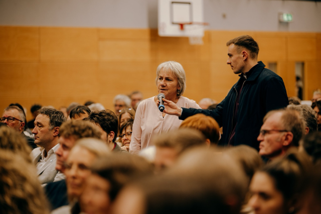 Eine Frau steht inmitten des Publikums und spricht in ein Mikrofon, das ihr ein Mann vor das Gesicht hält. © Staatsministerium BW | Ilkay Karakurt