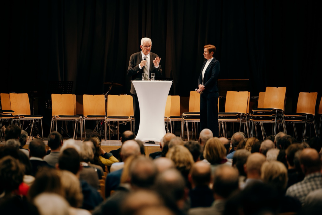 Sicht aus den hinteren Publikumsreihen auf die Bühne, wo Ministerpräsident steht und in ein Mikrofon spricht. Rechts neben ihm steht die Moderatorin. © Staatsministerium BW | Ilkay Karakurt