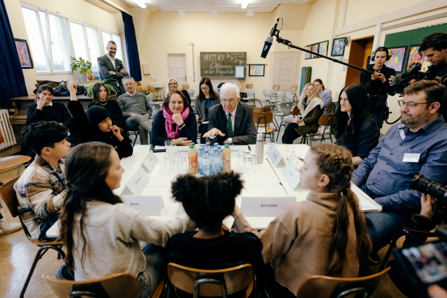 Ministerpräsident Kretschmann und Kultusministerin Schopper sitzen mit fünf Kindern und zwei Erwachsenen am Tisch und unterhalten sich. Im Hintergrund sieht man weitere Leute und eine Schultafel. Ein Mikro wird von einem Kamerateam in die Tischmitte gehalten.