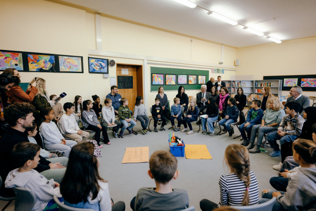 Ministerpräsident Kretschmann und Kultusministerin Schopper sitzen hinter einem großen Stuhlkreis, in dem Kinder und Erwachsene sitzen. Im Hintergrund halten Menschen Mikros und Smartphones hoch.