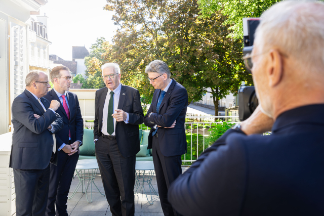 Ministerpräsident Kretschmann steht mit drei Männern in einem Halbkreis und unterhält sich. Alle tragen Anzug. Die Männer hören dem Ministerpräsidenten zu. Im Vordergrund ein Mann mit Kamera, der die Gruppe fotografiert.