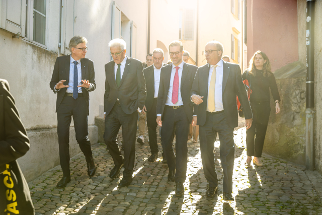 Ministerpräsident Kretschmann läuft in der Mitte von drei weiteren Männern über ein Kopfsteinpflaster. Er hört einem der Männer zu. Im Hintergrund sind weitere Menschen zu sehen, die ihnen hinterherlaufen. Die Sonne legt ein schönes Licht auf die Menschengruppe.
