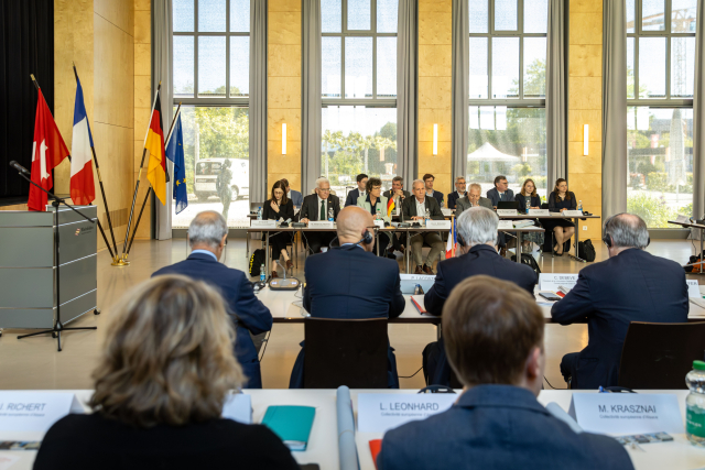 Ministerpräsident Kretschmann sitzt umringt von vielen Leuten in einem großen Saal. Im Hintergrund sind die Flaggen von Deutschland, der Schweiz und Frankreich zu sehen. Viele tragen Headsets.