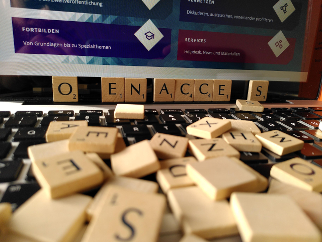 Das Bild zeigt Buchstabensteine auf einer Laptop-Tastatur, die sich zu "Open Access" zusammensetzen sollen, wobei das P und ein S noch fehlen. Im Hintergrund ist der Bildschirm eines Laptops sichtbar, der verschiedene Menüpunkte der Webseite open-access.network anzeigt. Im Vordergrund liegen auf der Tastatur mehrere Scrabble-artige Buchstabensteine, wobei die Buchstaben "O", "E", "N", "A", "C", "C", "E", "S" der Reihe nach am oberen Rand der Weitere Buchstabensteine sind verstreut auf der Tastatur.