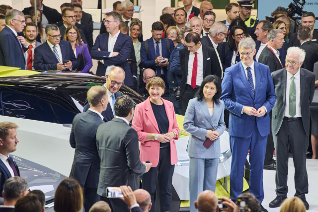 Ministerpräsident Kretschmann steht gemeinsam mit weiteren Funktionären aus Politik und Automobilwirtschaft auf einer Bühne. Im Hintergrund ist ein Auto zu sehen und eine größere Gruppe von Menschen.
