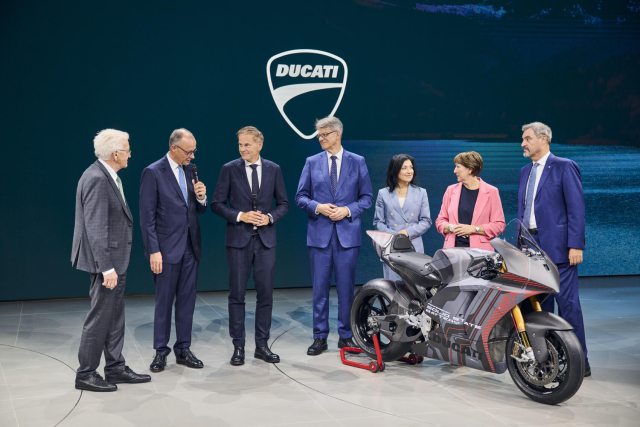 Ministerpräsident Kretschmann steht neben Bundeskanzler Merz und weiteren Funktionären aus Politik und Automobilwirtschaft auf einer Bühne. Er wendet sich dem Bundeskanzler zu, der in ein Mirko spricht. Vor ihnen steht ein Motorrad. Im Hintergrund eine blaue Wand, auf der "Ducati" aufgedruckt ist.