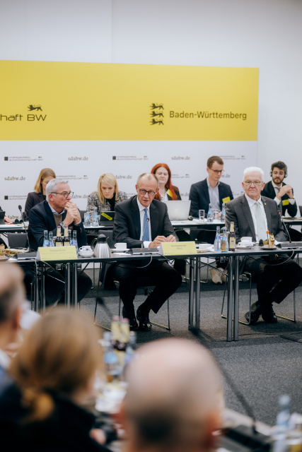 Bundeskanzler Merz sitzt zwischen Ministerpräsident Kretschmann und  dem stellvertretenden Ministerpräsidenten Strobl. Im Hintergrund sitzen weitere fünf Personen, außerdem ist eine gelb-weiße Wand mit der Aufschrift "Baden-Württemberg" zu sehen.